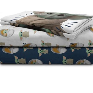 May include: A stack of three fitted bed sheets featuring the character Grogu from The Mandalorian. The top sheet has a large image of Grogu, while the other two sheets have repeating patterns of Grogu in a pod. The sheets are white and navy blue.