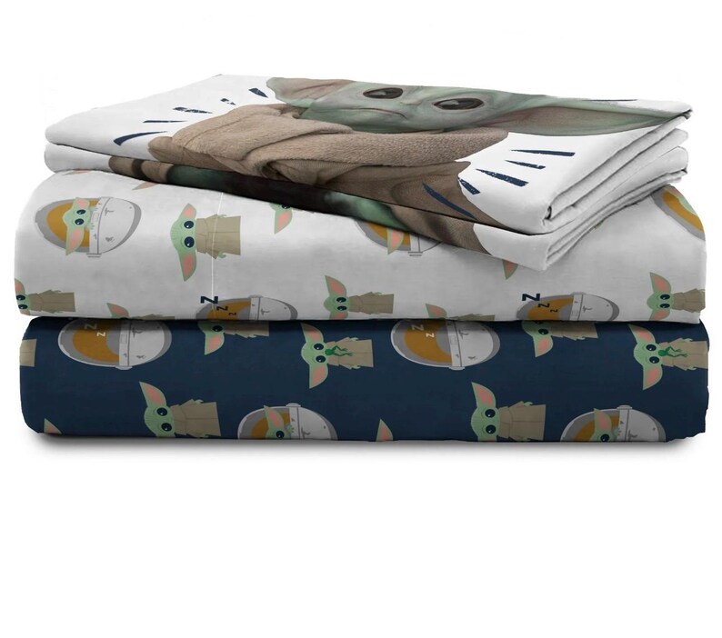 May include: A stack of three fitted bed sheets featuring the character Grogu from The Mandalorian. The top sheet has a large image of Grogu, while the other two sheets have repeating patterns of Grogu in a pod. The sheets are white and navy blue.