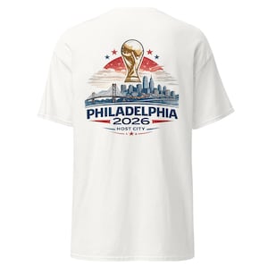 May include: White t-shirt featuring a graphic design for the 2026 Philadelphia Host City. The design includes a gold trophy, a cityscape, and the text "PHILADELPHIA 2026 HOST CITY" in blue and red.