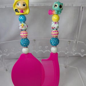 May include: Two pink hair combs with beaded handles. One handle features a yellow figurine with a microphone, the other a green chameleon. The handles have blue, floral, and pink beads.