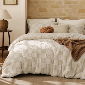 May include: A bed featuring a cream and beige checkered tufted duvet cover, two matching checkered pillow shams, two solid cream pillows, a round brown knot pillow, and a textured brown throw blanket draped across the foot of the bed.