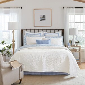 May include: A bed with a white quilted coverlet, a blue and white patterned bed skirt, and multiple pillows in shades of white and blue. The bed features a dark metal frame and is flanked by two floor lamps and a wooden side table.