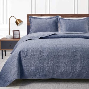 May include: A blue quilted bedspread and two matching pillow shams. The bedding features a repeating floral pattern. The bed is made up with the quilt and pillows, ready for use. The bedroom has a nightstand and lamp.