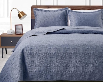 May include: A blue quilted bedspread and two matching pillow shams. The bedding features a repeating floral pattern. The bed is made up with the quilt and pillows, ready for use. The bedroom has a nightstand and lamp.