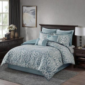 May include: A blue and white damask patterned comforter set on a bed. The set includes a comforter, bed skirt, and several pillows. The bedroom has a dark wood headboard, nightstand, and dresser.