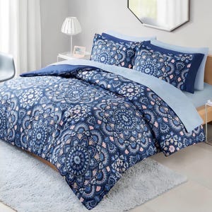 May include: A blue and white patterned bedspread with matching pillowcases. The bedding features a floral mandala design in shades of blue and white, with accents of pink. The bed is made up with a light blue sheet and a dark blue comforter.
