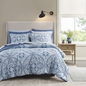 May include: A bed with a light beige headboard is covered with a blue and white patterned comforter and matching pillowcases. The pattern features a circular design. The room has a neutral color scheme with a wooden nightstand and a window.