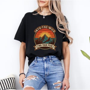 Puede incluir: Una camiseta negra de manga corta con un gráfico circular que muestra una cordillera, pinos y una puesta de sol en tonos naranja, amarillo y azul verdoso. El texto "INTO THE WILD" y "FIND YOUR PEAK" está impreso en una fuente llamativa.