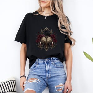 以下が含まれることがあります： 黒の半袖Tシャツ。中央には開いた本、その周りにダークレッドのバラ、2本の灯ったキャンドル、黒い鳥が描かれたグラフィックプリントがあり、全体が精巧なゴールド調のフィリグリー模様で縁取られています。