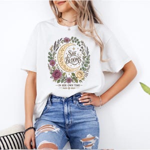 Pode incluir: Uma t-shirt branca de manga curta com um gráfico de uma lua crescente rodeada por uma coroa de flores. O texto "SHE BLOOMS" está centrado dentro da lua e "IN HER OWN TIME" está impresso abaixo do design floral em tinta preta.
