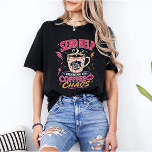 May include: A black short-sleeve t-shirt featuring a graphic of a coffee mug with steam and the text "SEND HELP", "FRESH COFFEE", "RUNNING ON COFFEE AND CHAOS". The design includes pink lightning bolts, yellow stars, and decorative lines in pink and yellow.