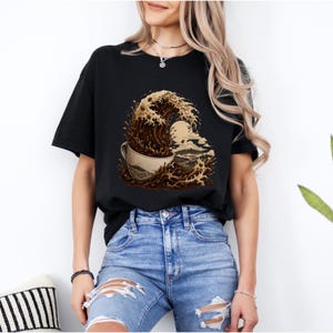 Puede incluir: Camiseta negra con un diseño gráfico único de una taza de café con una ola de café y una escena de montaña. El diseño es en tonos marrones y beige. Una prenda casual para el día a día.