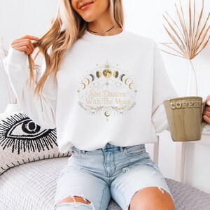 Pode incluir: Uma camisola branca de manga comprida com um gráfico central das fases da lua num padrão circular. O texto "She Dances With The Moon" está impresso ao centro numa fonte dourada, rodeado por delicados desenhos botânicos lineares.