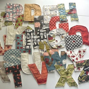 May include: A collection of fabric alphabet letters featuring various patterns including farm animals, cherries, gingham checks, and vintage-style prints. Each letter is individually stitched with a decorative pinked edge, showcasing a diverse array of colors and motifs.