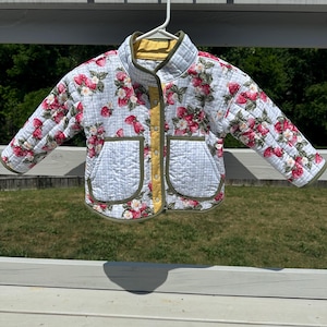 Könnte beinhalten: Eine gesteppte Jacke mit weiß-hellblauem Vichy-Muster und einem Aufdruck aus roten Erdbeeren und weißen Blüten. Die Jacke hat einen Stehkragen, lange Ärmel, zwei aufgesetzte Vordertaschen und eine gelbe Knopfleiste mit olivgrüner Einfassung.