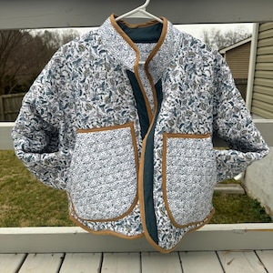 May include: A quilted jacket with a white background and a floral pattern in shades of blue, green, and brown. The jacket has two front pockets, a dark green interior, and brown trim. It is hanging on a white hanger.