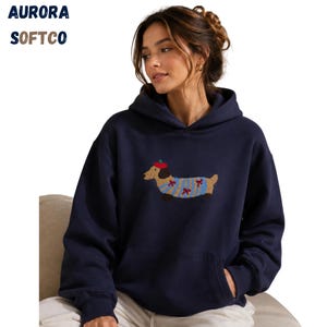 K&ouml;nnte beinhalten: Ein marineblauer Kapuzenpullover mit einer Stickerei eines beigen Dackels, der ein rotes Barett und einen blau gestreiften Pullover mit kleinen roten Schleifen tr&auml;gt. Der Text AURORA SOFTCO ist oben links in blauen und braunen Buchstaben gedruckt.