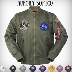 Op de afbeelding: Olijfgroene bomberjack met ritssluiting, geribde manchetten en kraag. De jas heeft een Apollo 11-patch, een NASA-patch en een kleine Amerikaanse vlag. De tekst "REMOVE BEFORE FLIGHT" staat op een rood label.
