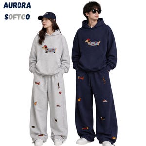 K&ouml;nnte beinhalten: Ein hellgrauer Kapuzenpullover mit passender Jogginghose, bestickt mit Dackeln und roten Herzen, sowie ein dunkelblauer Kapuzenpullover mit passender Hose und &auml;hnlichen Dackelstickereien. Der Text lautet AURORA SOFTCO.