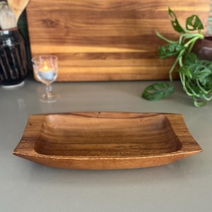 May include: A rectangular wooden serving tray with a smooth, polished finish and a deep, concave center. The tray features a natural brown wood grain pattern with slightly flared, elevated edges on the left and right sides.