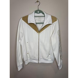 May include: A white zip-up track jacket featuring gold-colored panels across the chest and shoulders, a high collar, and a gold zipper pull. The left sleeve displays the word Adidas in gold lettering with the trefoil logo underneath the text.
