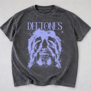Op de afbeelding: Donkergrijs t-shirt met de bandnaam "Deftones" in lichtpaars boven een lichtpaarse afbeelding van een gezicht met handen over de ogen. Het shirt heeft een ronde hals en korte mouwen.