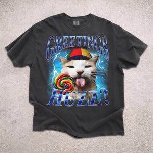 Op de afbeelding: Donkergrijs t-shirt met een kat die een kleurrijke hoed draagt en een regenboog lolly vasthoudt. Het shirt heeft de tekst "GREETINGS HUZZ?" in blauw, met een bliksemachtergrond. De kat steekt zijn tong uit.