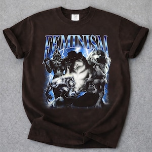 Puede incluir: Camiseta marrón oscuro con un diseño gráfico. El diseño presenta la palabra "FEMINISM" en una fuente azul con forma de rayo y varias criaturas parecidas a lobos en tonos grises y blancos.
