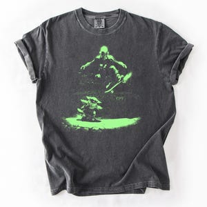 May include: Dark gray t-shirt with a neon green graphic of two figures skateboarding. The top figure is mid-air, while the bottom figure is on the ground. The shirt has a crew neck and short sleeves.