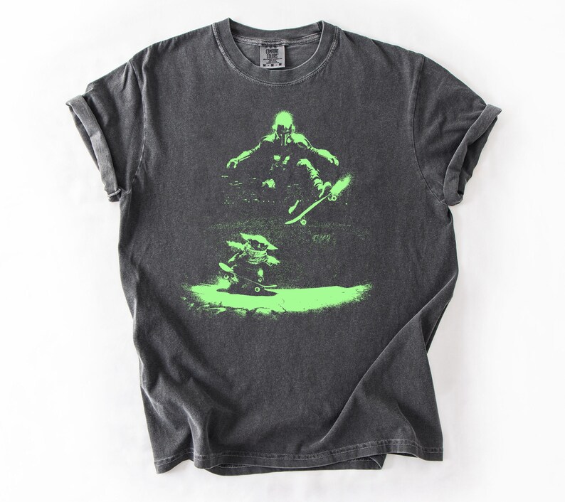 May include: Dark gray t-shirt with a neon green graphic of two figures skateboarding. The top figure is mid-air, while the bottom figure is on the ground. The shirt has a crew neck and short sleeves.
