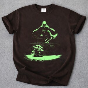 May include: A brown t-shirt featuring a neon green graphic of two figures skateboarding. One figure is in the air, while the other is on the ground. The shirt has rolled-up sleeves and a crew neck.