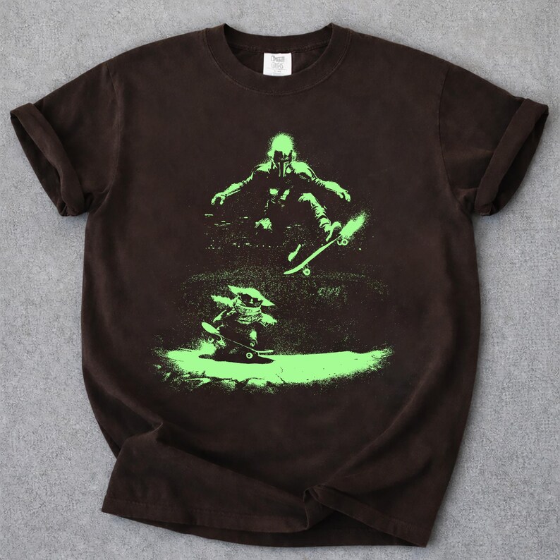 May include: A brown t-shirt featuring a neon green graphic of two figures skateboarding. One figure is in the air, while the other is on the ground. The shirt has rolled-up sleeves and a crew neck.