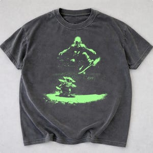 May include: Dark gray t-shirt featuring a neon green graphic of two figures skateboarding. The top figure is a person in a helmet and the bottom figure is a small creature. The t-shirt has a crew neck and short sleeves.