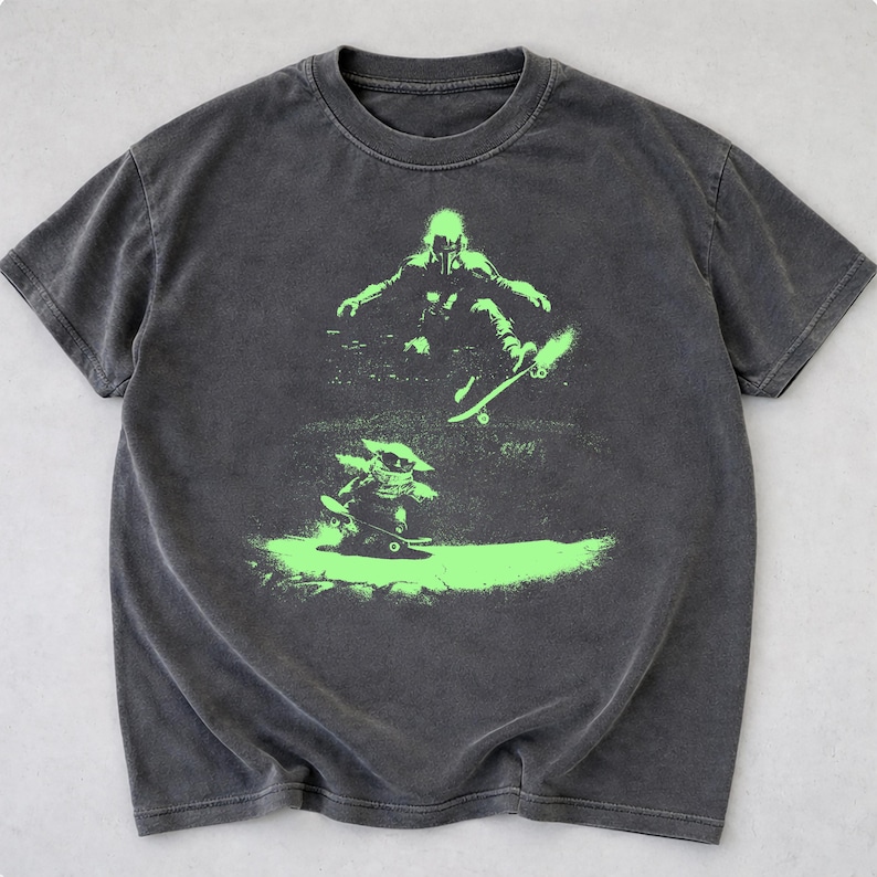 May include: Dark gray t-shirt featuring a neon green graphic of two figures skateboarding. The top figure is a person in a helmet and the bottom figure is a small creature. The t-shirt has a crew neck and short sleeves.