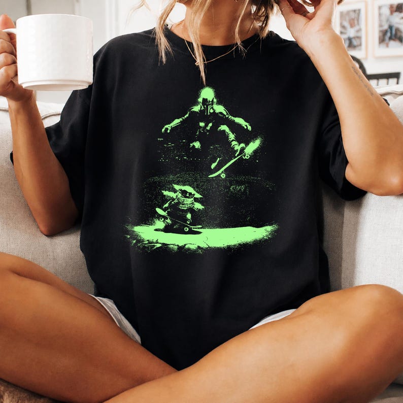 May include: Black t-shirt featuring a neon green graphic of two figures skateboarding. The top figure is mid-air, performing a trick, while the bottom figure is on the ground. The design has a spray-paint effect.
