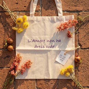 May include: A beige canvas tote bag featuring the purple embroidered text L'Amore non ha dress code. The bag is decorated with yellow flowers, orange floral sprigs, and small brown fruits, and includes a white tag with handwritten text.