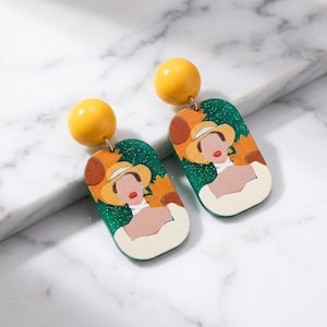 Può includere: Un paio di orecchini con borchie gialle e pendenti rettangolari. I pendenti raffigurano una donna con un cappello su uno sfondo verde glitterato. Gli orecchini sono un mix di giallo, verde, marrone e crema.