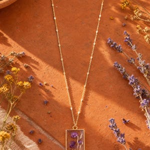 Puede incluir: Un collar de tono dorado con un colgante rectangular que encierra flores moradas prensadas. La cadena es delicada e incluye pequeños detalles geométricos dorados espaciados uniformemente, asegurada con un cierre de mosquetón.