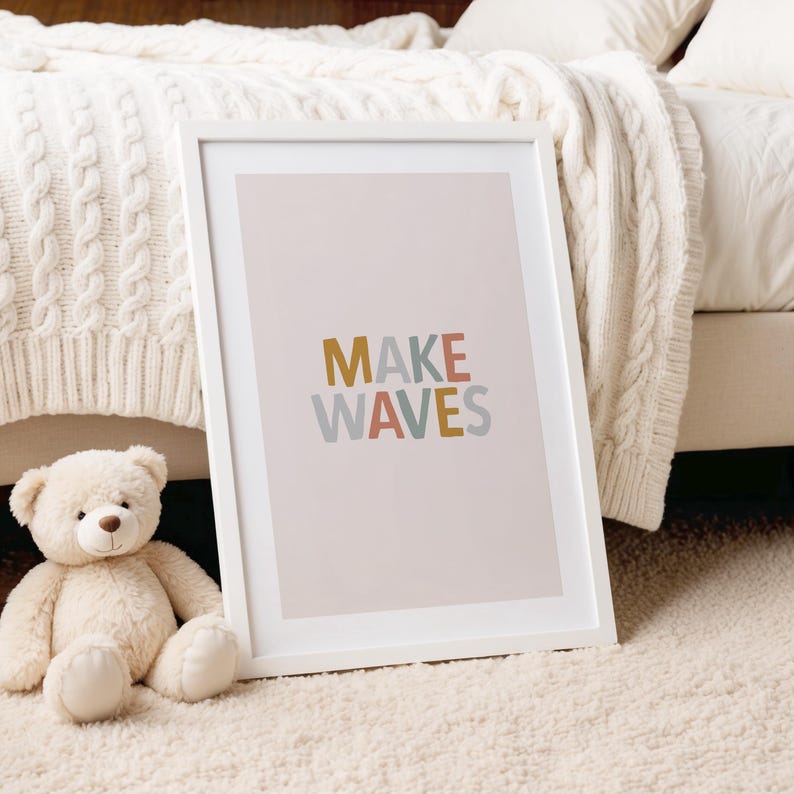May include: A white framed print with the words "MAKE WAVES" in colorful letters. The print is leaning against a bed with a white cable knit blanket. A stuffed teddy bear sits on the floor in front of the print.