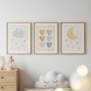 Pode incluir: Três impressões de arte emolduradas com um tema de berçário. Uma impressão apresenta uma nuvem sorridente, outra tem corações coloridos e a terceira mostra uma lua crescente sorridente. As impressões estão em molduras de madeira clara e são exibidas em uma parede branca.