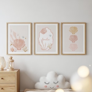May include: Three framed art prints with a coastal theme. Each print features a light beige background with watercolour illustrations of seashells and aquatic plants in shades of pink, peach, and beige. One print includes the name "Amelia".