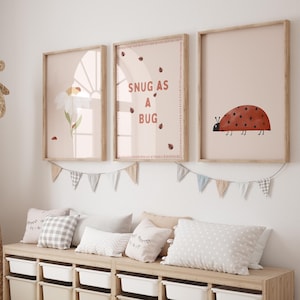 Peut inclure: Trois impressions d'art encadrées avec des cadres en bois clair sont accrochées au-dessus d'un banc de rangement en bois. Les impressions présentent une marguerite, le texte "SNUG AS A BUG" et une illustration de coccinelle. Une guirlande de petits drapeaux et des coussins décorent l'espace.