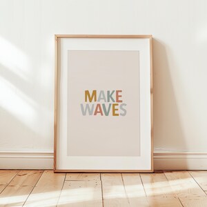 May include: A framed art print with the words "MAKE WAVES" in mustard yellow, coral, and blue-gray lettering. The print has a light pink background and is set in a wooden frame. The artwork is displayed against a white wall and wooden floor.