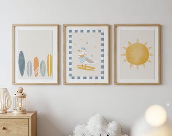 May include: Three framed art prints with a beach theme. The prints feature a row of surfboards, a seagull on a surfboard, and a yellow sun. The artwork is in soft pastel colors, with a light wood frame.