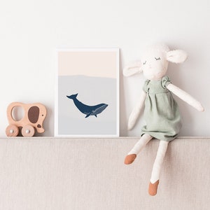May include: A white framed print with a blue whale illustration on a light blue and pink background. A stuffed white lamb wearing a green dress sits on a beige couch.