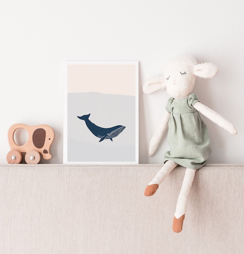 May include: A white framed print with a blue whale illustration on a light blue and pink background. A stuffed white lamb wearing a green dress sits on a beige couch.