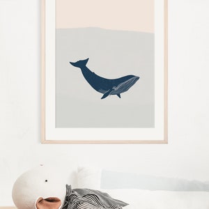 May include: A framed print of a blue whale silhouette swimming in a light blue and peach colored ocean. The print is hanging on a white wall above a white bed with a striped pillow and a white stuffed animal.