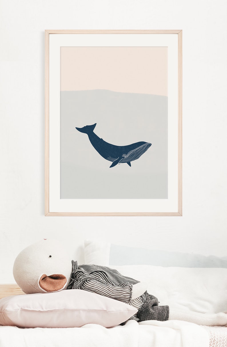 May include: A framed print of a blue whale silhouette swimming in a light blue and peach colored ocean. The print is hanging on a white wall above a white bed with a striped pillow and a white stuffed animal.