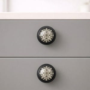 May include: Two black cabinet knobs with a decorative cream and black floral design. The knobs are round and set against a gray cabinet drawer. The knobs are a decorative element for furniture.