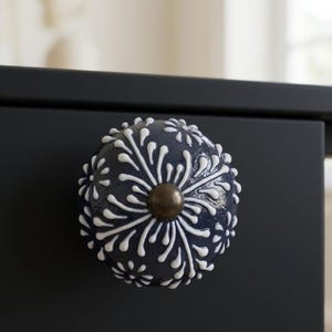 May include: A navy blue cabinet knob with a white floral design and a bronze center. The decorative knob is round and appears to be ceramic. The knob is attached to a dark blue cabinet.
